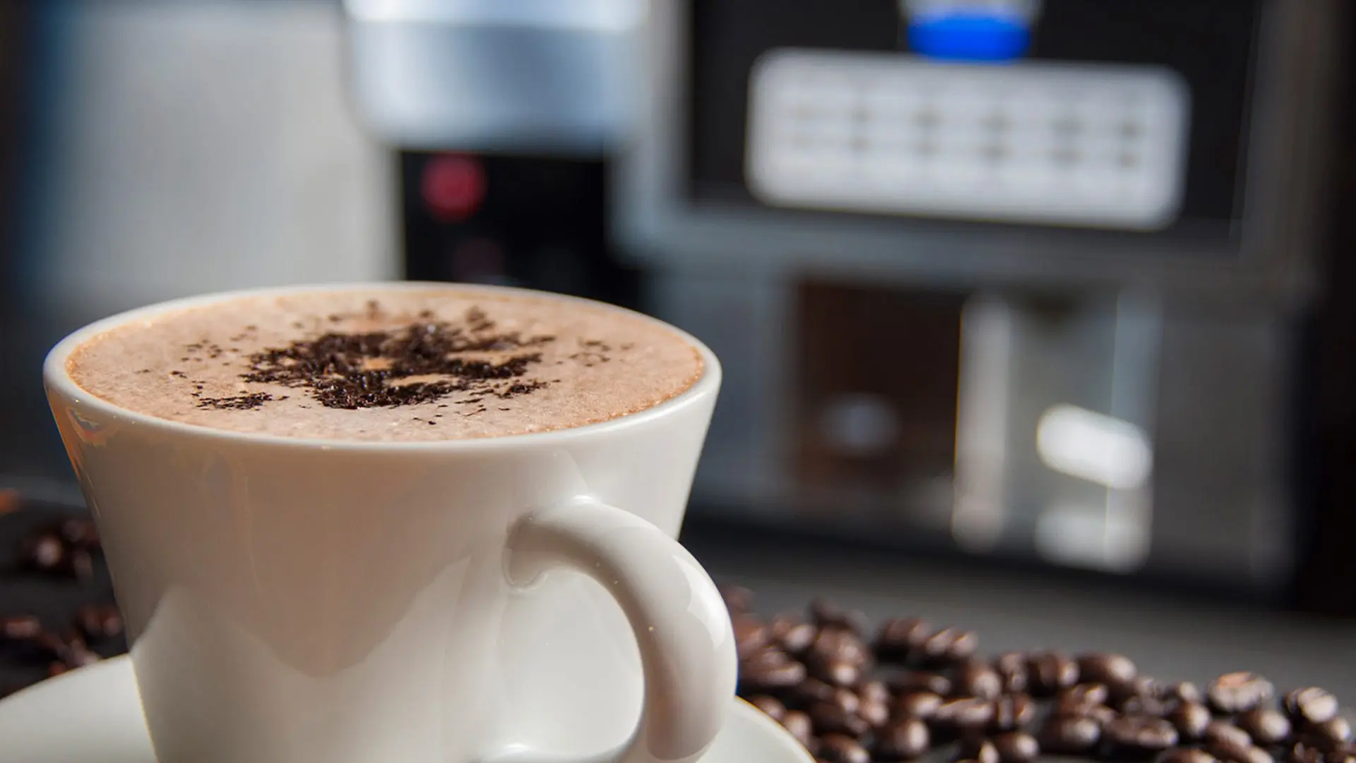 Cup of cappuccino topped with chocolate shavings beside coffee beans, espresso machine in the background