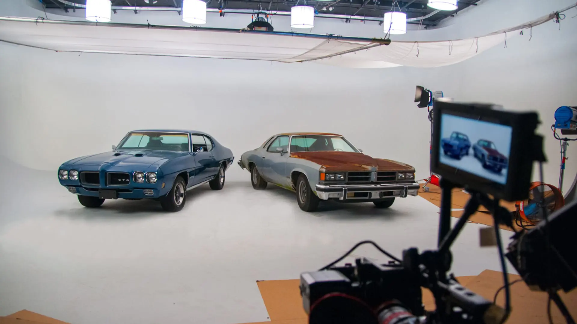 Two classic cars, one blue and one with a rusted hood, on a white cyc stage with a camera and monitor in the foreground