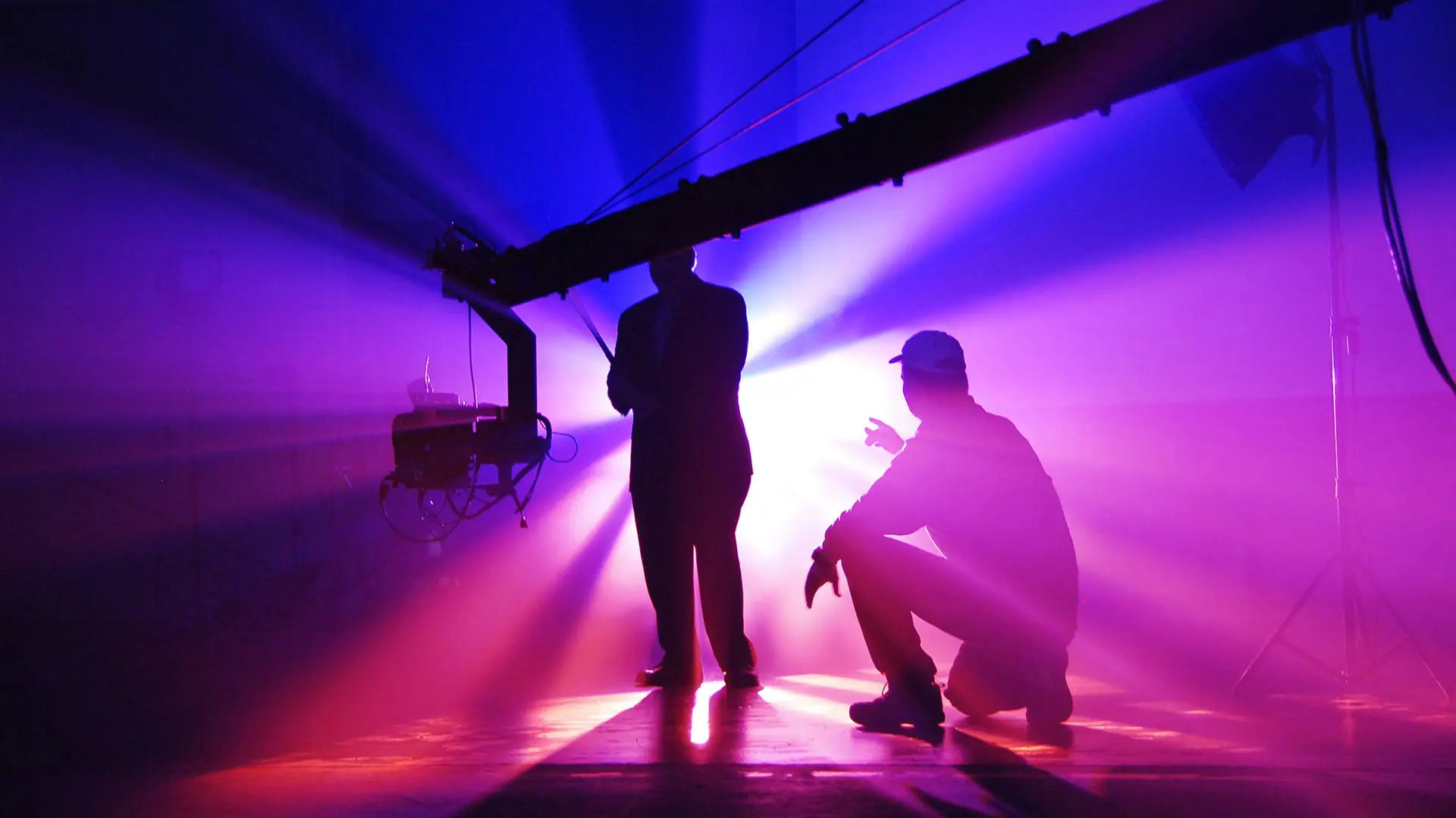 Two crew members silhouetted under a camera jib, backlit by purple and pink light beams in a studio