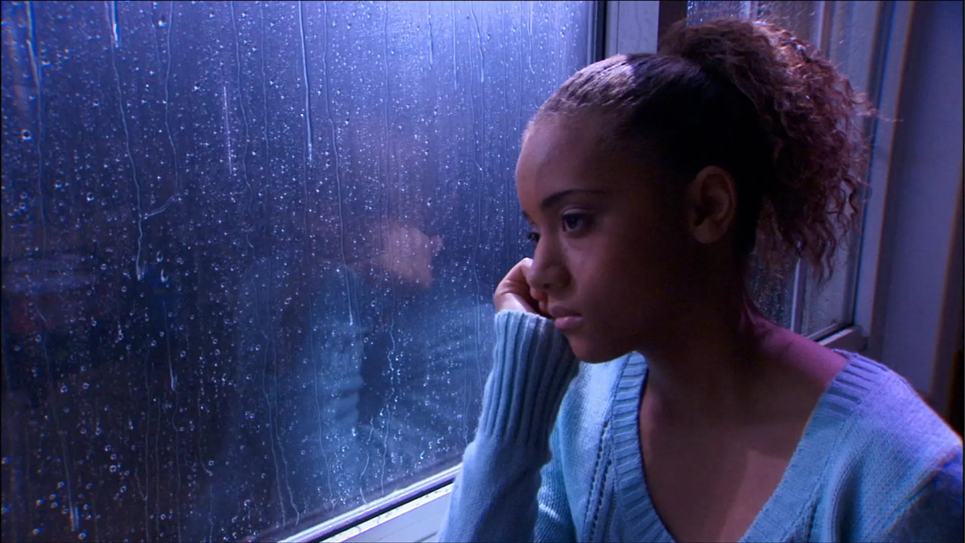 Young woman in a blue sweater looking out a rain-covered window