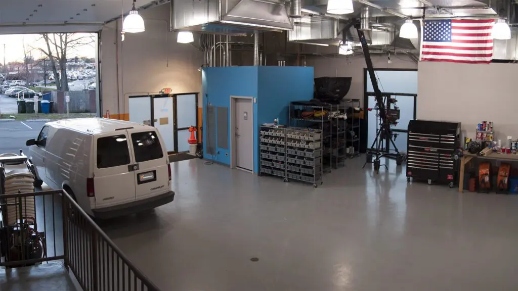 Truck bay with a white van parked indoors, parts carts, a camera crane, tool chest and American flag