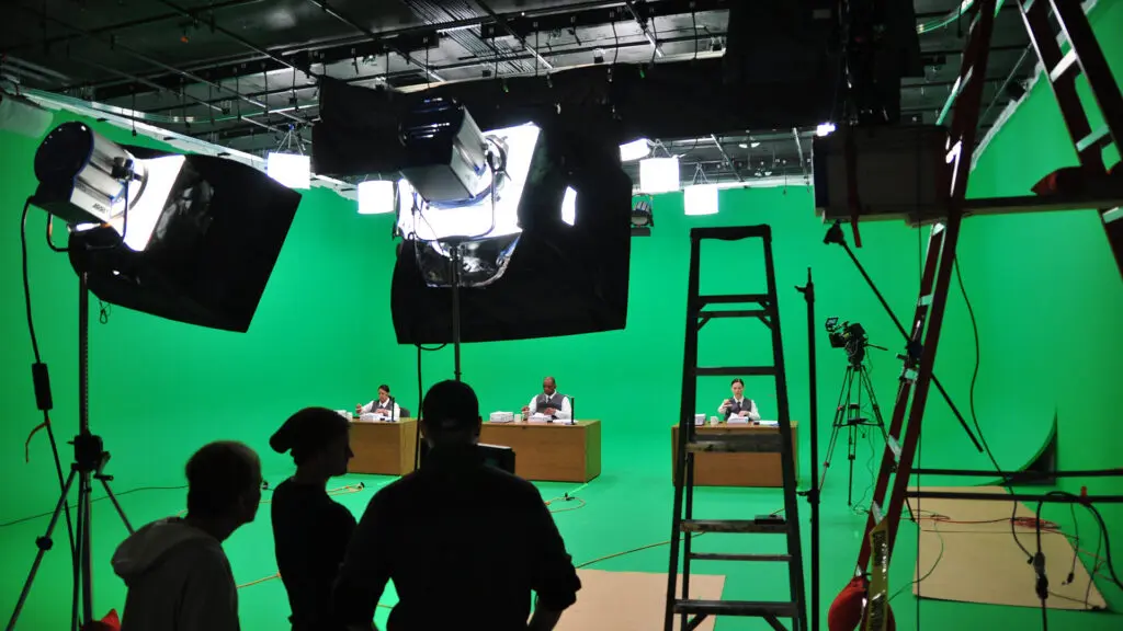 Three people seated at desks on a green screen set, with crew silhouetted in the foreground