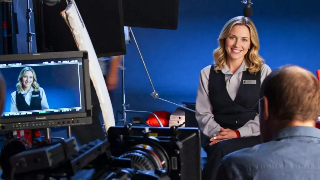 Smiling woman in a vest seated for an interview against a blue backdrop, with a monitor showing her shot
