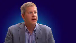 Man in a blazer seated for an interview against a dark blue background